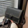A black leather mini cross-body bag with gold studs and a brown patterned adjustable strap is shown next to a window blind.