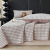 A plaid quilted bedspread in soft pink and beige tones is draped over a dark headboard with a teddy bear pillow.