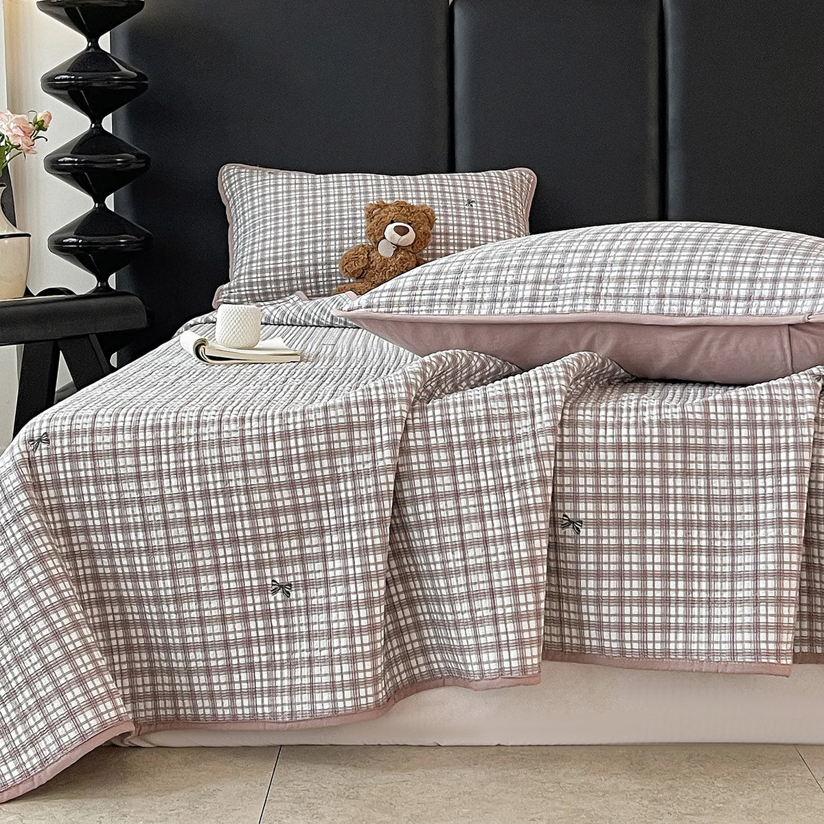 A plaid quilted bedspread in soft pink and beige tones is draped over a dark headboard with a teddy bear pillow.