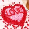 A heart shape made of red and pink silk rose petals scattered on a white bed sheet.