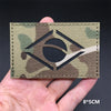 A close-up of a rectangular tactical patch featuring a black eye symbol on a camouflage background being held by a hand.
