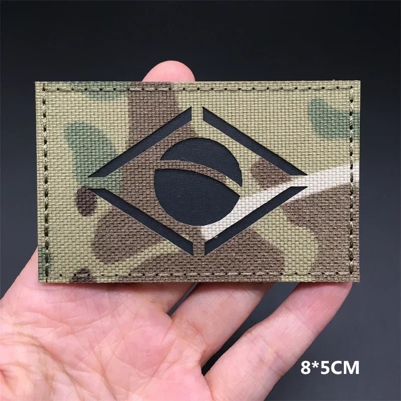 A close-up of a rectangular tactical patch featuring a black eye symbol on a camouflage background being held by a hand.