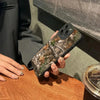 A hand holds a phone with a forest camouflage silicone case against a gray background.