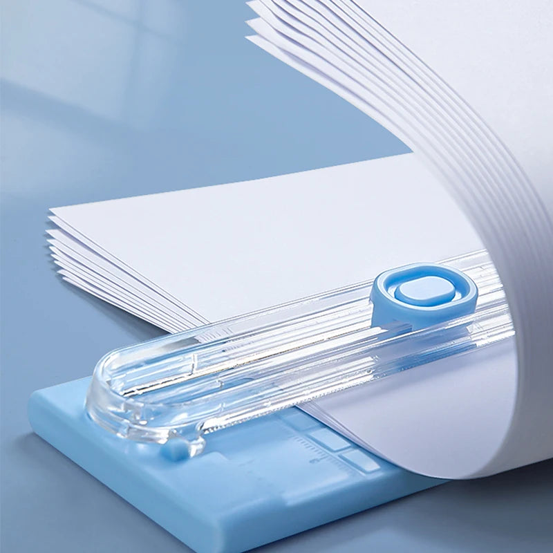 A close-up view of a portable manual paper cutter trimmer with a clear plastic blade and white paper, set against a light blu