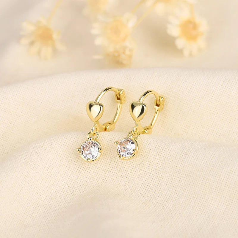 Pendientes de aro dorados con piedras de circonita cúbica transparente, exhibidos en tela crema.