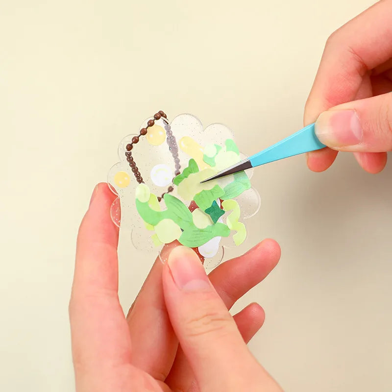Hands use blue tweezers to place macaron color craft elements onto a sheet for scrapbooking