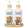 A white soap dispenser bottle with a wooden pump and label showing a Highland cow face on the front, sitting on a wooden tray
