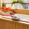 A hand is placing a gray cleaning brush with a white scouring head into a white kitchen sink.