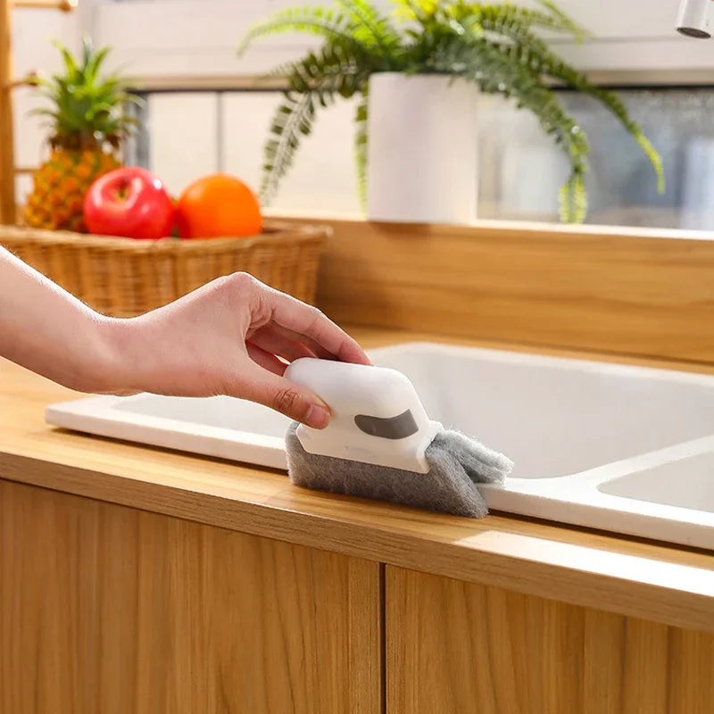 A hand is placing a gray cleaning brush with a white scouring head into a white kitchen sink.