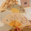 A light wood folding fan with a floral design is displayed on a white cloth.