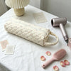 A quilted cotton hair dryer storage bag with a handle resting on a white tablecloth next to a pink comb and some rose petals.