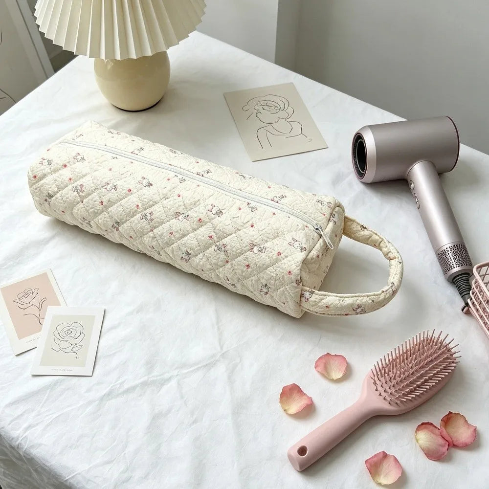 A quilted cotton hair dryer storage bag with a handle resting on a white tablecloth next to a pink comb and some rose petals.