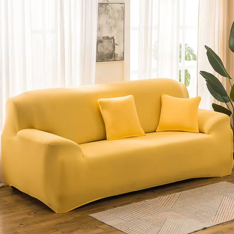 A mustard yellow elastic stretch sofa cover for a 3-seater sofa with two accent pillows, placed in a bright living room.