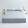 A light blue cooling ice-silk fitted sheet with two pillows is displayed on a white surface next to a modern lamp.