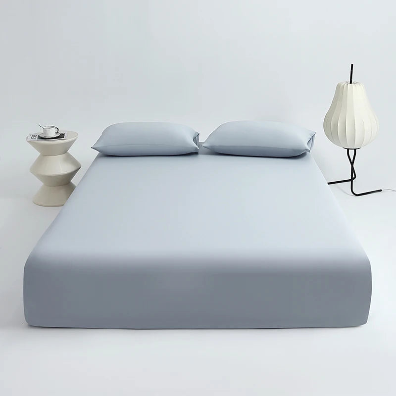 A light blue cooling ice-silk fitted sheet with two pillows is displayed on a white surface next to a modern lamp.