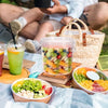 A transparent reusable storage container with a handle and leakproof lid filled with colorful fruit slices is shown on a picn
