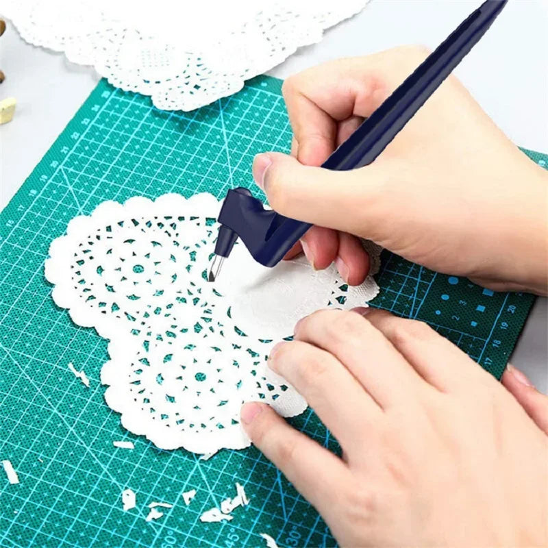A hand uses a dark blue precision craft knife to cut intricate lace patterns on white paper on a green cutting mat.