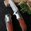 A folding pocket knife with a stainless steel blade and a walnut wood handle rests on a dark surface next to a piece of rock.