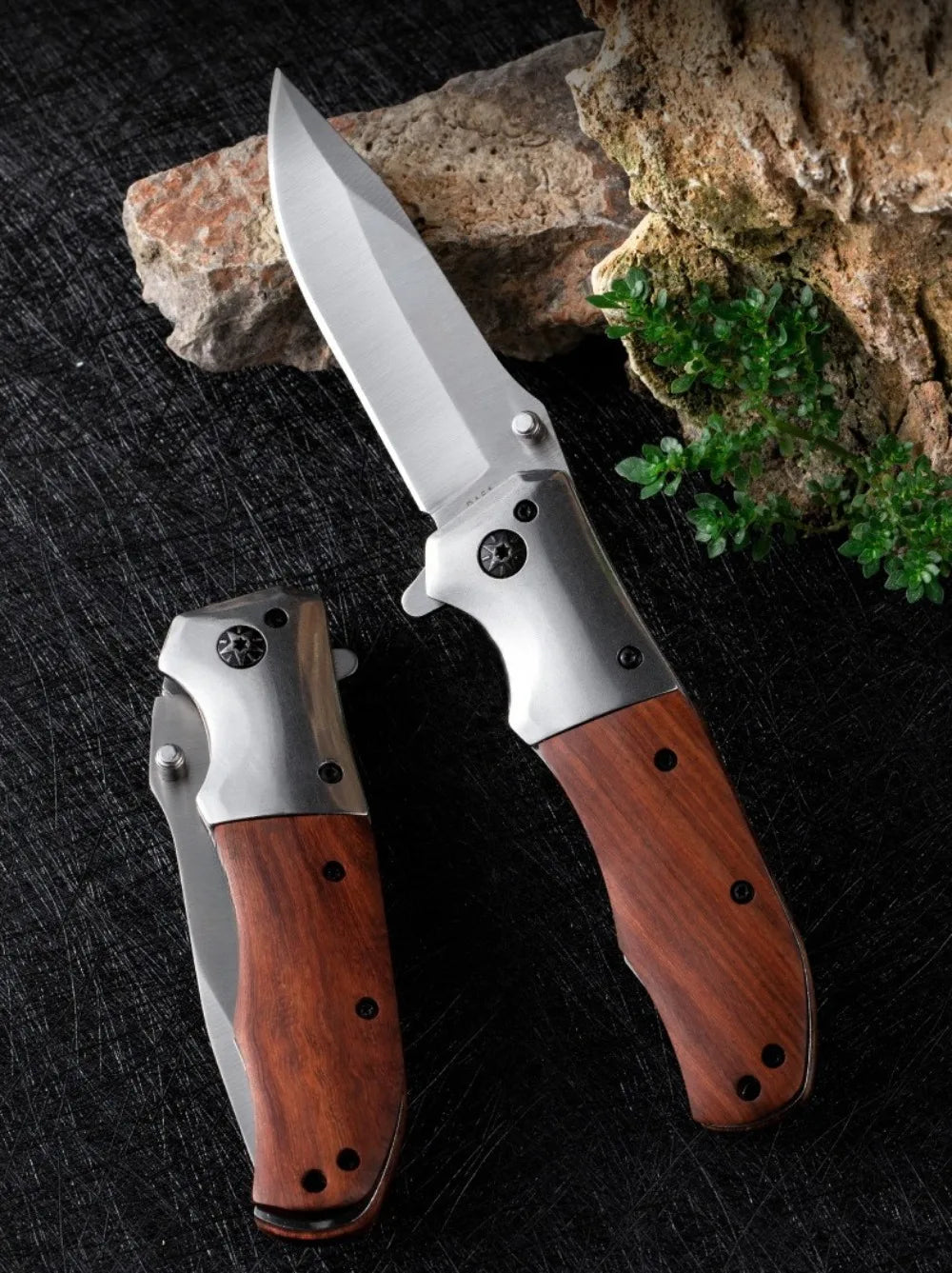 A folding pocket knife with a stainless steel blade and a walnut wood handle rests on a dark surface next to a piece of rock.
