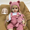 A lifelike silicone baby doll wearing a pink knitted outfit and a matching hat, sitting on a brown textured surface.