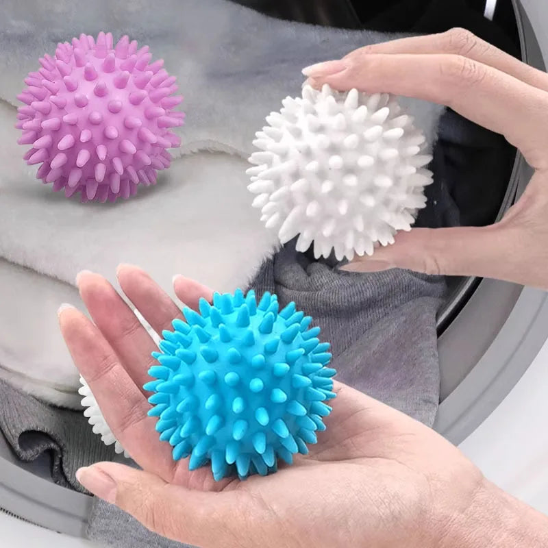 Three reusable laundry balls in pink, white, and blue are held in a hand against a gray fabric background.