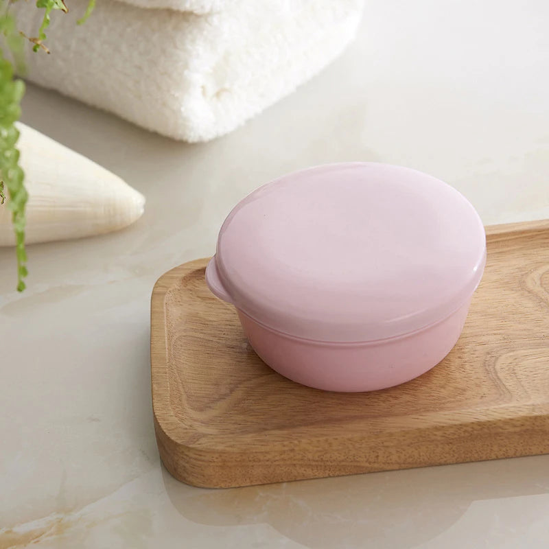 A pink sealed portable soap box with a drainage cover rests on a light wooden cutting board next to a white towel.