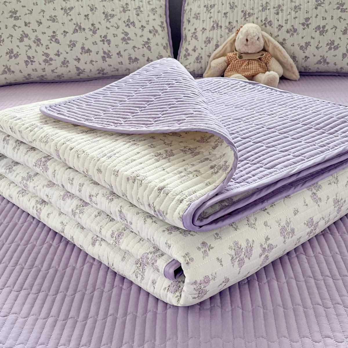 A lavender quilted bedspread with a floral pattern is folded on a purple surface next to a stuffed animal.
