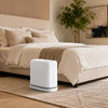 A modern white touchless trash can with auto bagging sits on a light wood floor in a bedroom setting.