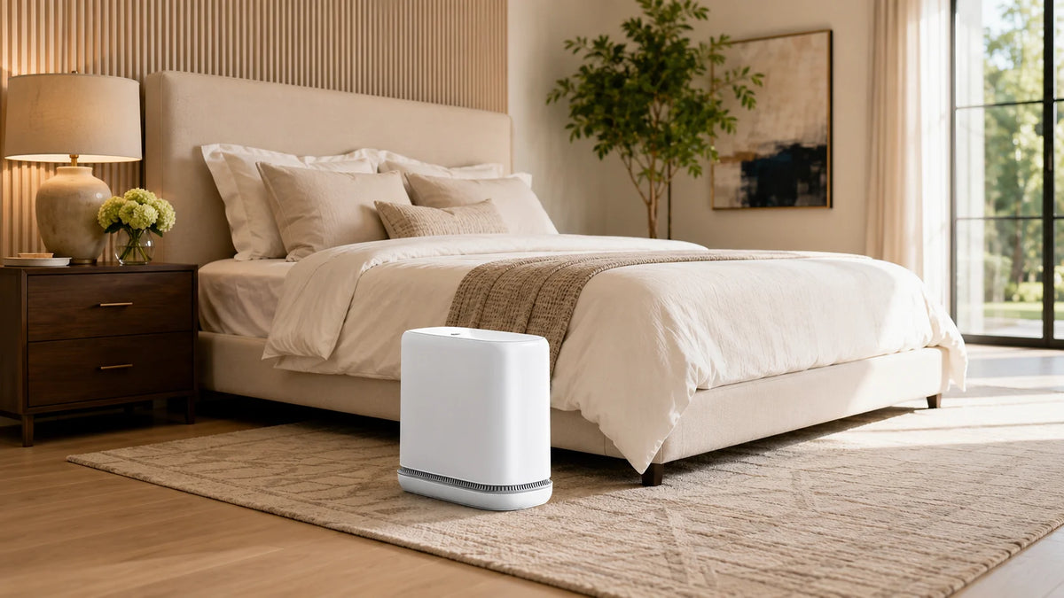 A modern white touchless trash can with auto bagging sits on a light wood floor in a bedroom setting.