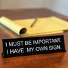 A black desk sign with white text that reads I MUST BE IMPORTANT I HAVE MY OWN SIGN resting on a wooden surface.