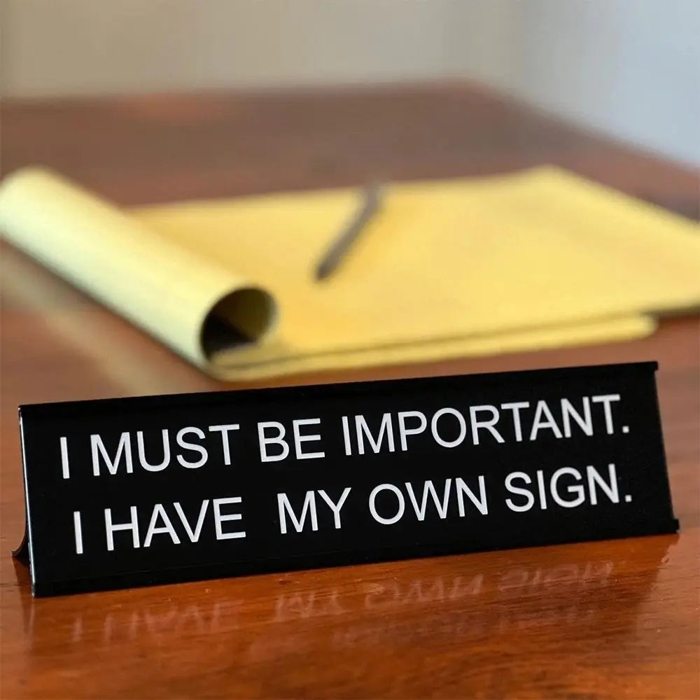 A black desk sign with white text that reads I MUST BE IMPORTANT I HAVE MY OWN SIGN resting on a wooden surface.