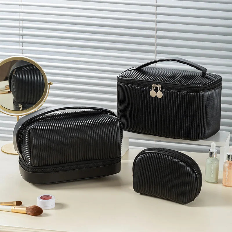 A set of three black PU leather makeup bags displayed on a light surface next to a mirror and cosmetic products.