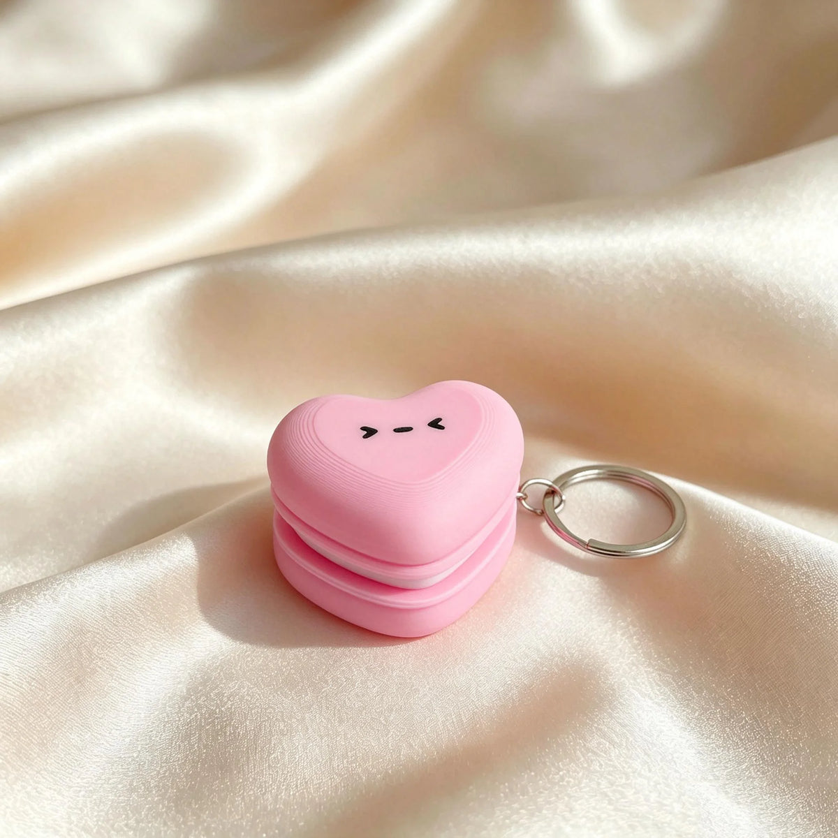 A pink heart-shaped fidget keychain with a small cartoon face is resting on silky beige fabric.