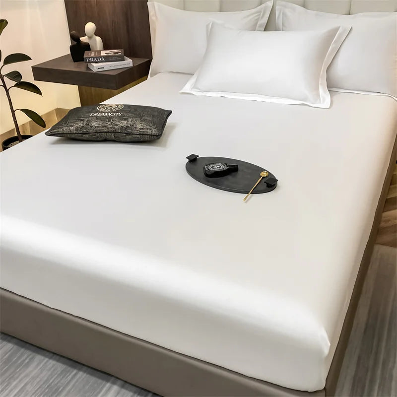 Un juego de sábanas blancas ajustadas se muestra en una cama, con almohadas y un pequeño accesorio negro en la sábana.