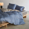 A luxury mulberry silk duvet cover set in blue and gray tones displayed on a bed with pillows and a bedside lamp.
