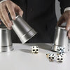 A hand is holding a silver tin while several white and black gothic skull dice are displayed on a white surface.