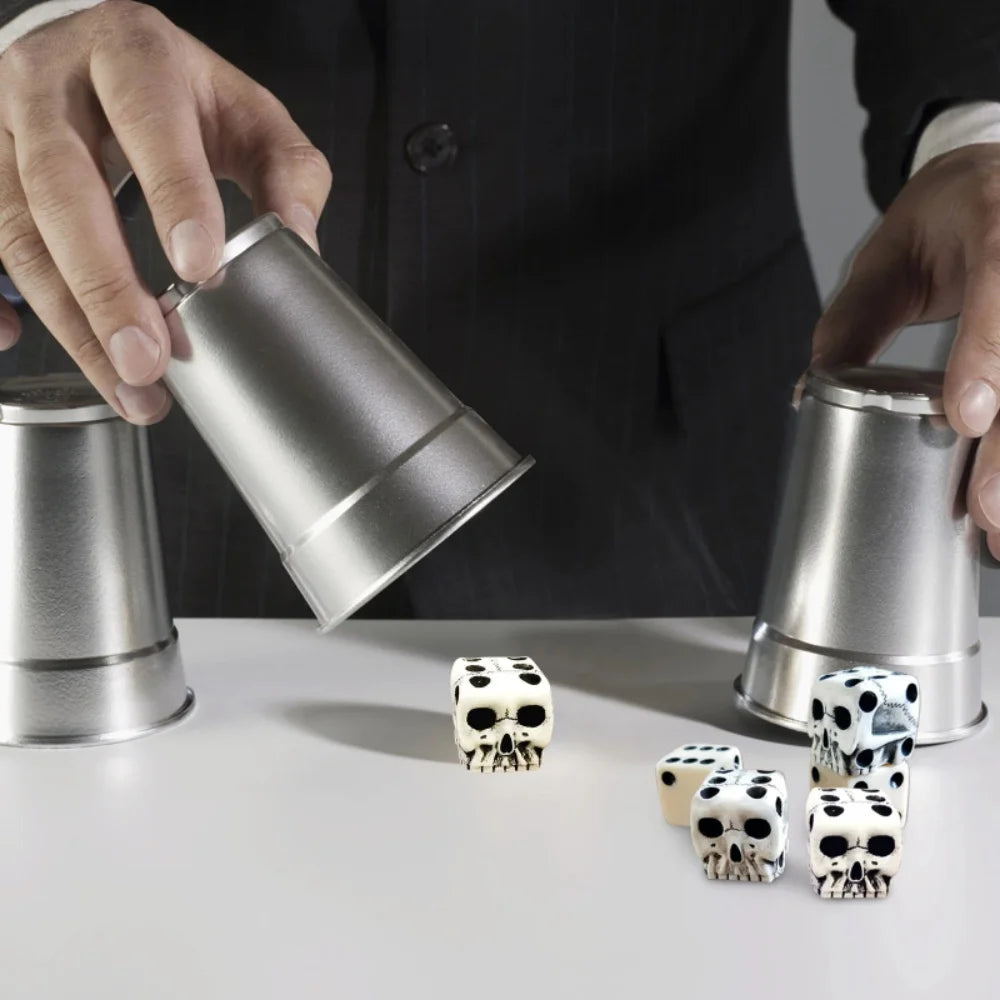 A hand is holding a silver tin while several white and black gothic skull dice are displayed on a white surface.