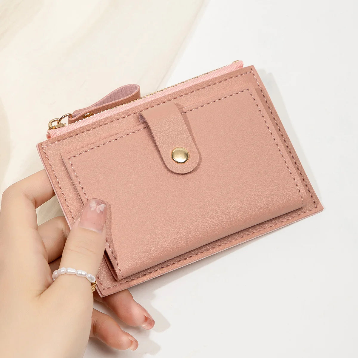 A hand holds a pink women's slim PU leather card holder wallet with a gold zipper and stud closure.