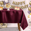 A burgundy satin tablecloth is set for a celebration with small cakes and gold balloons in the background.