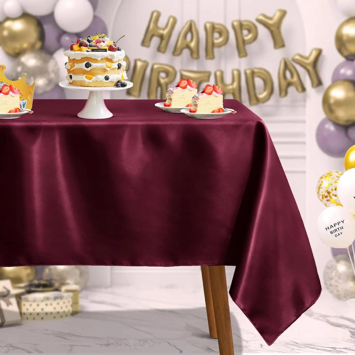 A burgundy satin tablecloth is set for a celebration with small cakes and gold balloons in the background.