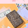 Leopardmuster-Budgetplaner-Notizbuch neben US-Dollar und Lineal auf orangem Hintergrund.