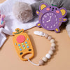 A silicone teether toy shaped like a yellow pineapple and a purple clock with a wooden bead bracelet on a soft fabric backgro
