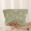 A hand holds a light green floral corduroy makeup bag with a zipper on a white lace surface.
