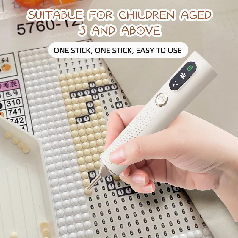 A hand holds a white electric diamond art pen with a vacuum suction feature, demonstrating its use on a grid surface.