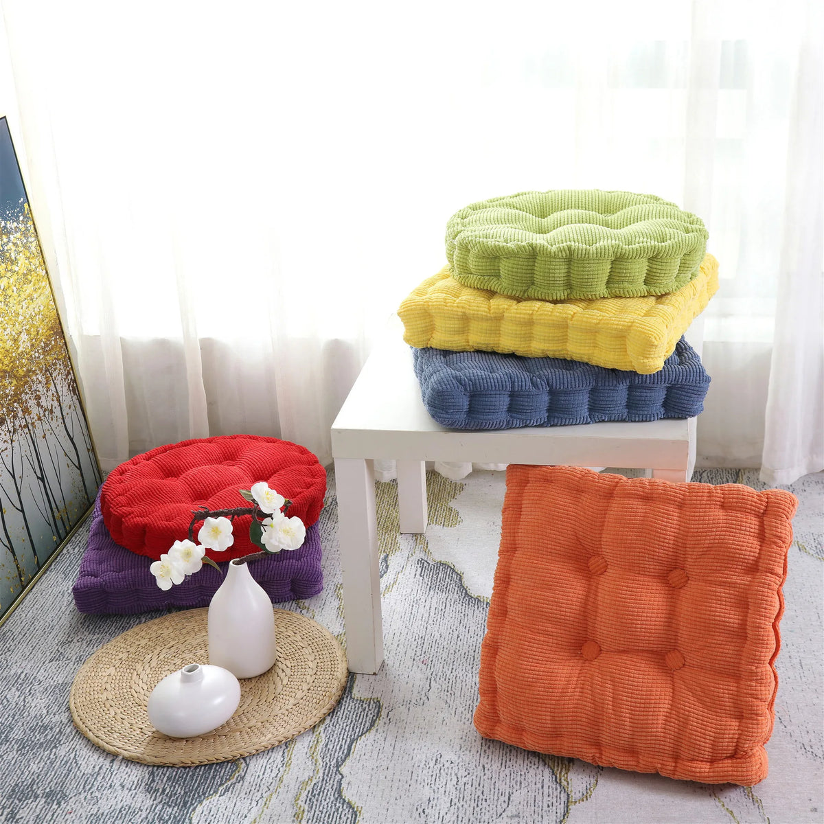A collection of thick corduroy floor cushions in various shades of green, yellow, and blue are displayed on a white table.