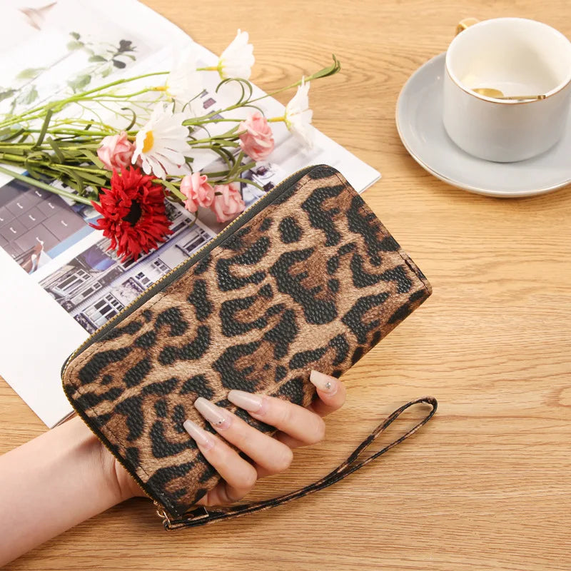 A leopard print retro long wallet is held by a hand on a wooden table next to a cup and some flowers.