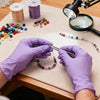 Hands in purple nitrile gloves are stringing beads onto a string under a magnifying glass.