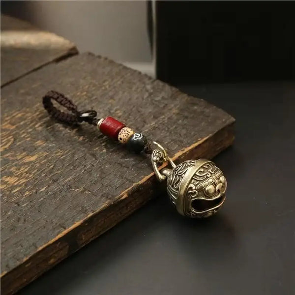 A vintage alloy bell wind chime keychain with a dark cord and an ornate metal bell resting on a wooden surface.