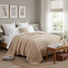 A beige pure cotton muslin throw blanket draped over a white bed with framed art on the wall.