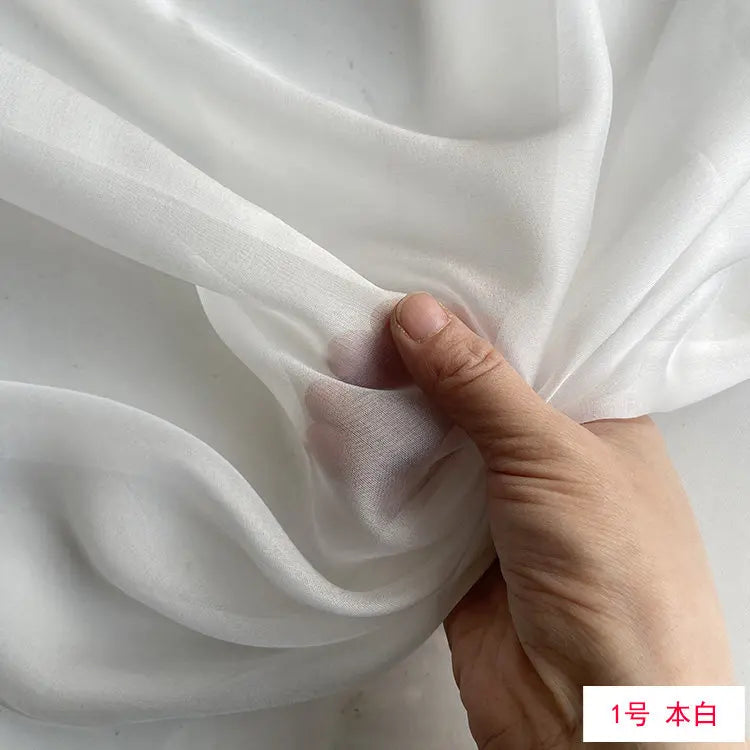 A hand is gently folding a piece of white, lightweight, sheer mulberry silk chiffon fabric.